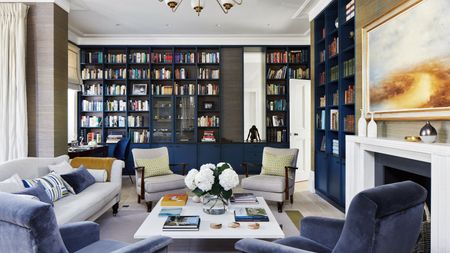 Living room with grey walls, dark blue fitted bookcase and display units, grey and blue sofa and armchairs around a white coffee table beside the fireplace.