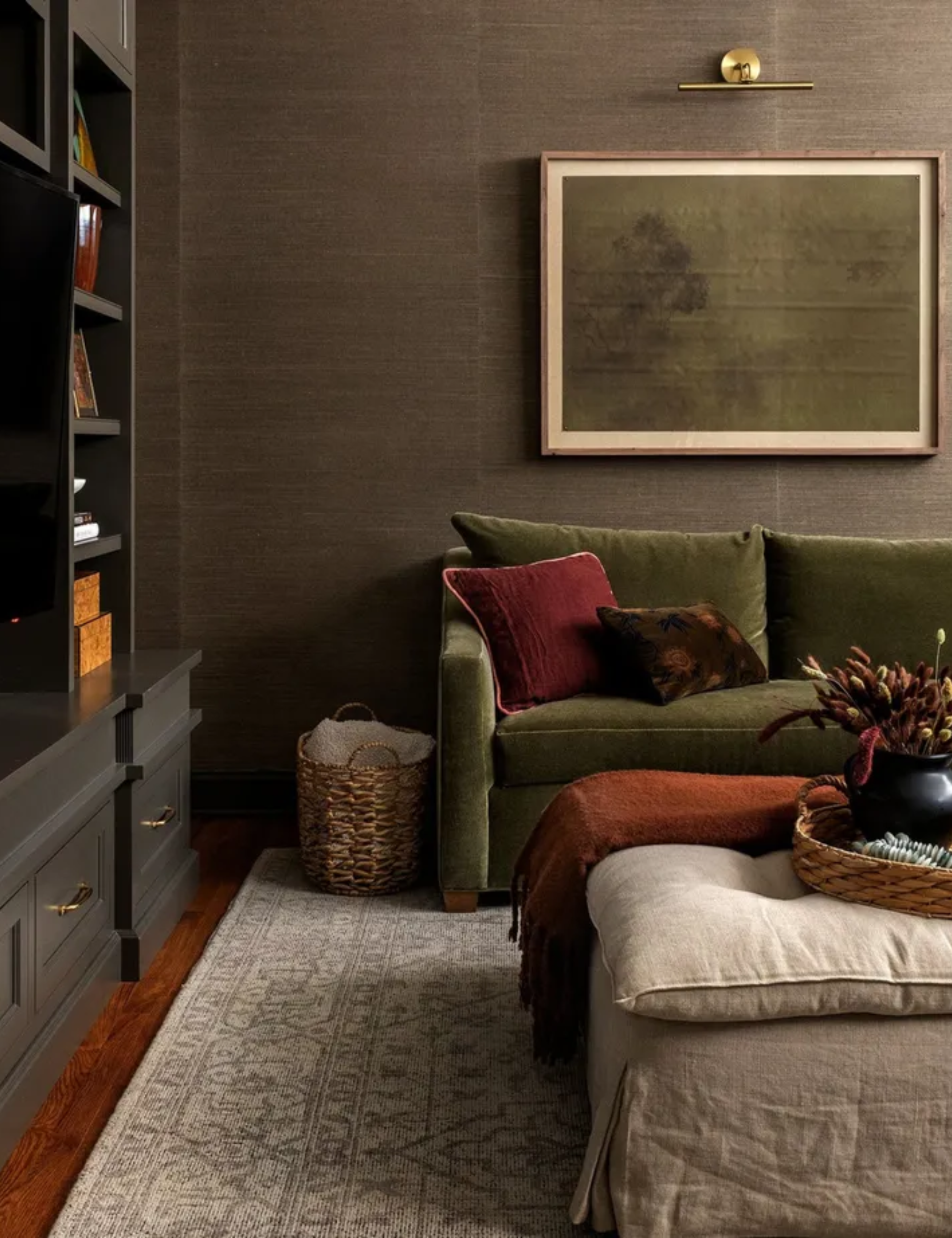 Dark living room with patterned rug, green sofa with cushions, framed minimalist painting, partial black bookshelf to the left, footstool, wooden floors.