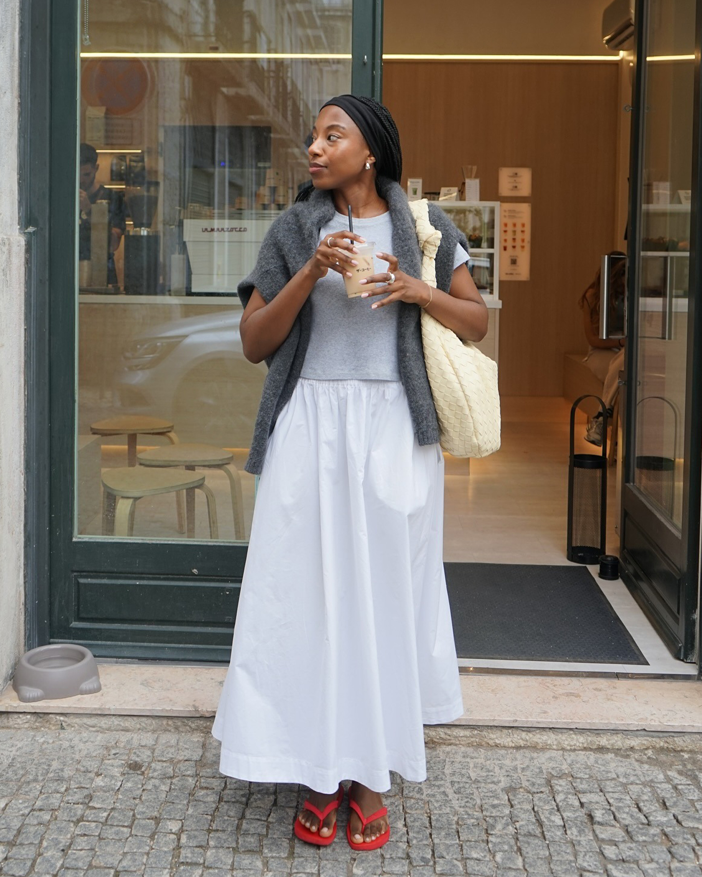British style influencer Taffy Msipa stands outside a cafe with an iced coffee wearing a gray t-shirt, gray sweater on her shoulders, butter-yellow woven shoulder bag, white full skirt, and red flat flip-flop sandals