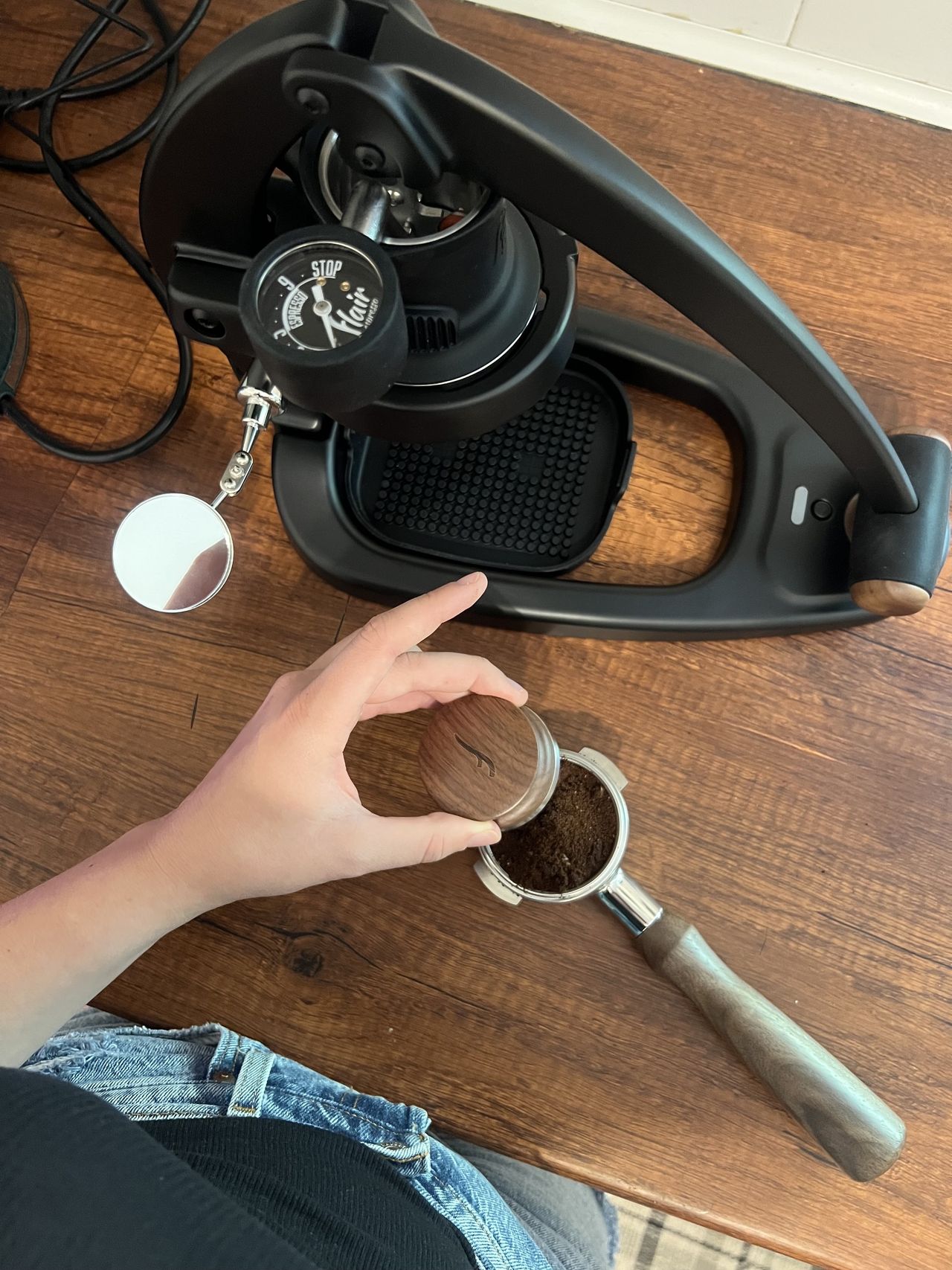 A wooden portafilter with grounds in it next to a black and wood manual coffee machine sitting on a wooden countertop in a kitchen with white tiles on the wall.