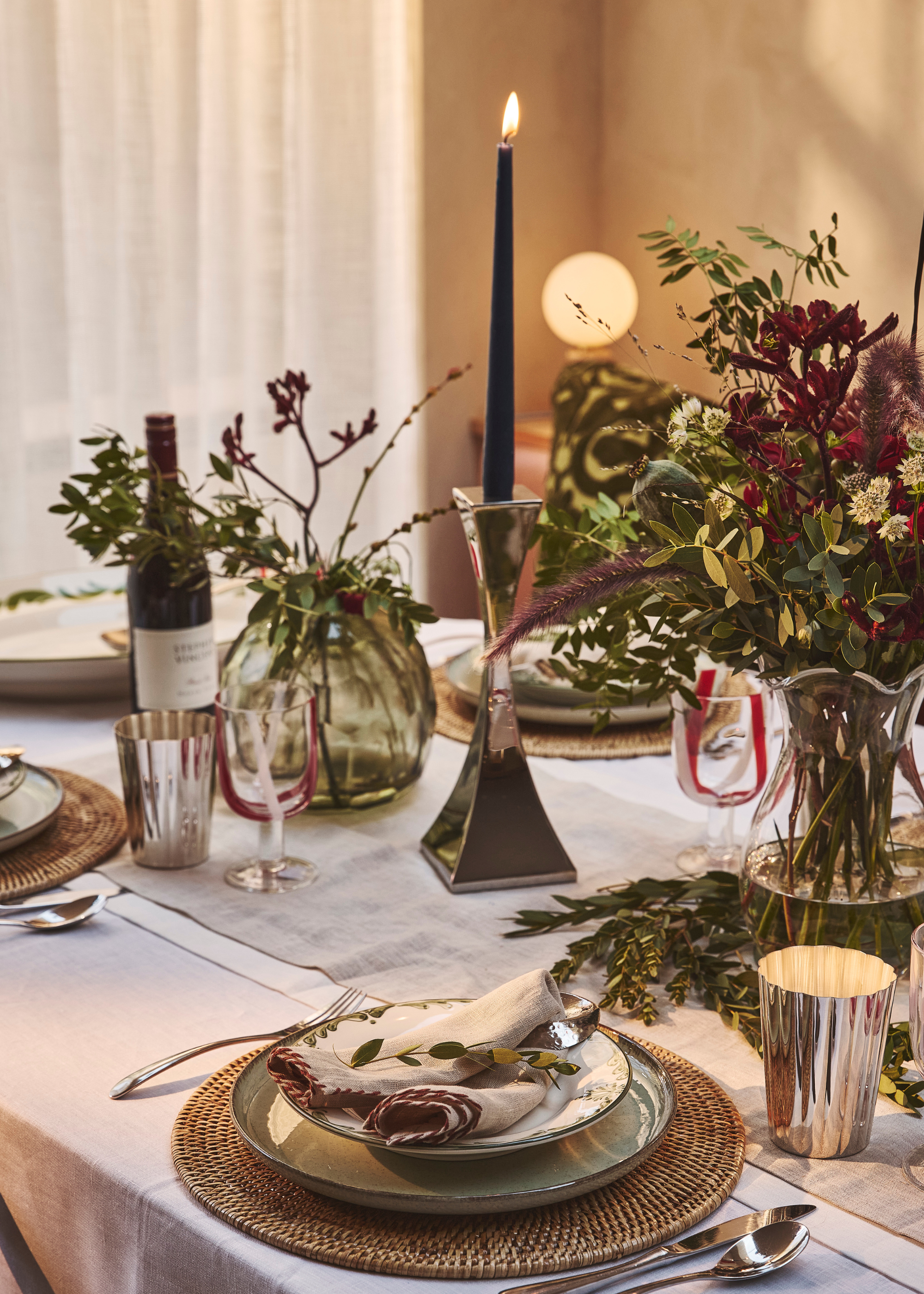A table set and decorated for Christmas with fresh florals and greenery, a back lit taper candle in the center in a sculptural chrome candelstick holder with ratan charger plates contrasting against the white linen tablecloth