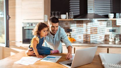 An astonished-looking toddler watches her smiling Dad work on his laptop at the kitchen counter.