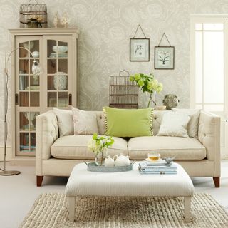 Neutral country-style living room with cream sofa and upholstered footstool and neutral wallpaper with a nature-inspired design