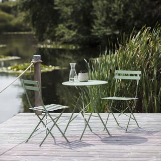 Sage green bistro table and chairs on decking next to lake.