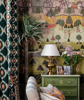 Maximalist corner of a living room with patterned curtains, artistic, painted mural style wallpaper, green plant and cabinet, striped lounge chairs, brass table lamp, colorful vase of flowers