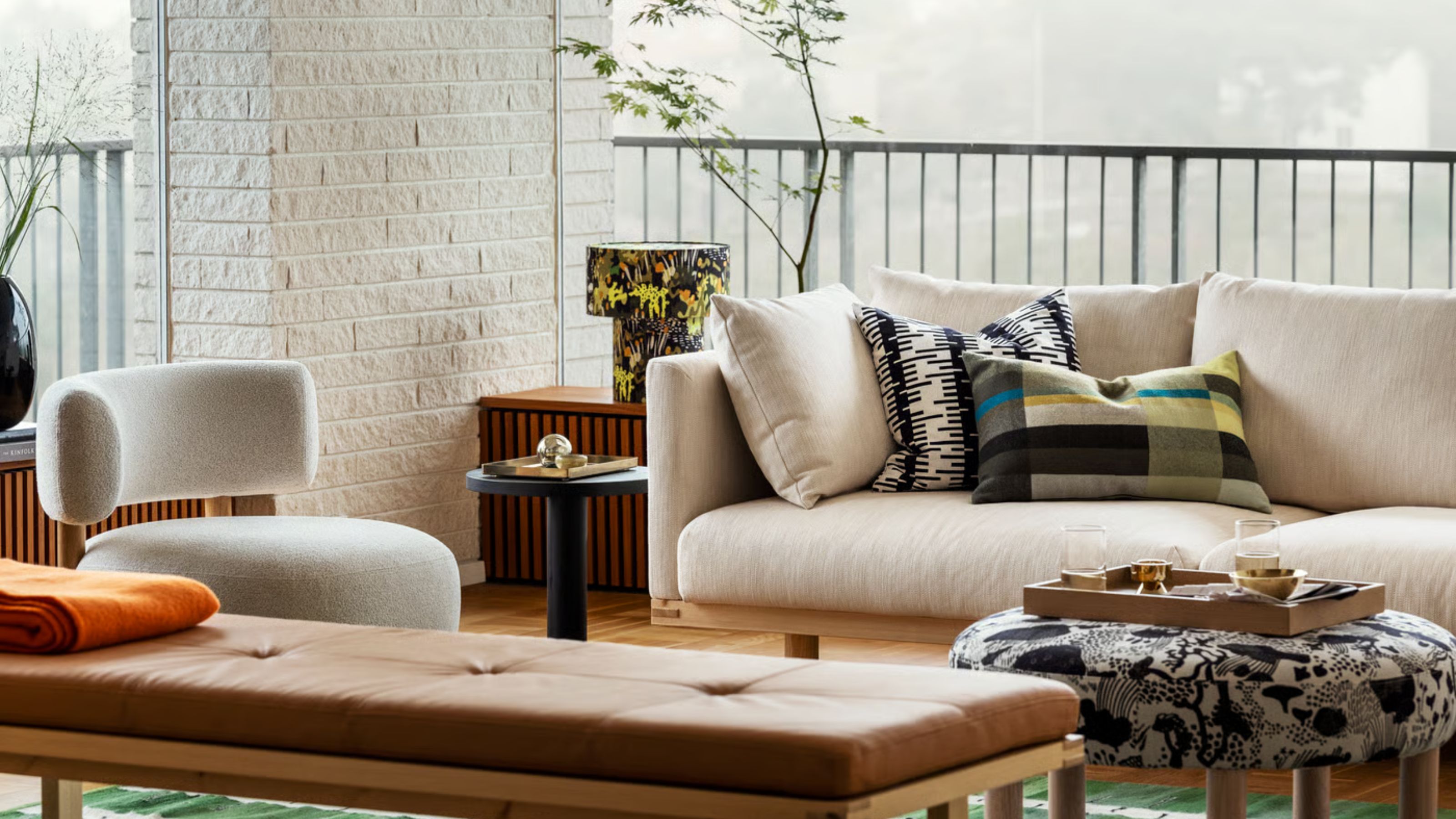 outdoor living area with white ikea stockholm sofa, bench seat, side table with patterned lamp, white boucle armchair, patterned upholstered ottoman with a tray with decor in it