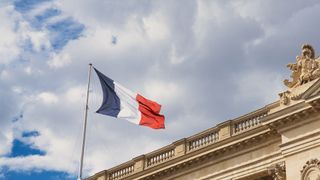 French flag pictured on top of a government building against a clear sky backdrop.