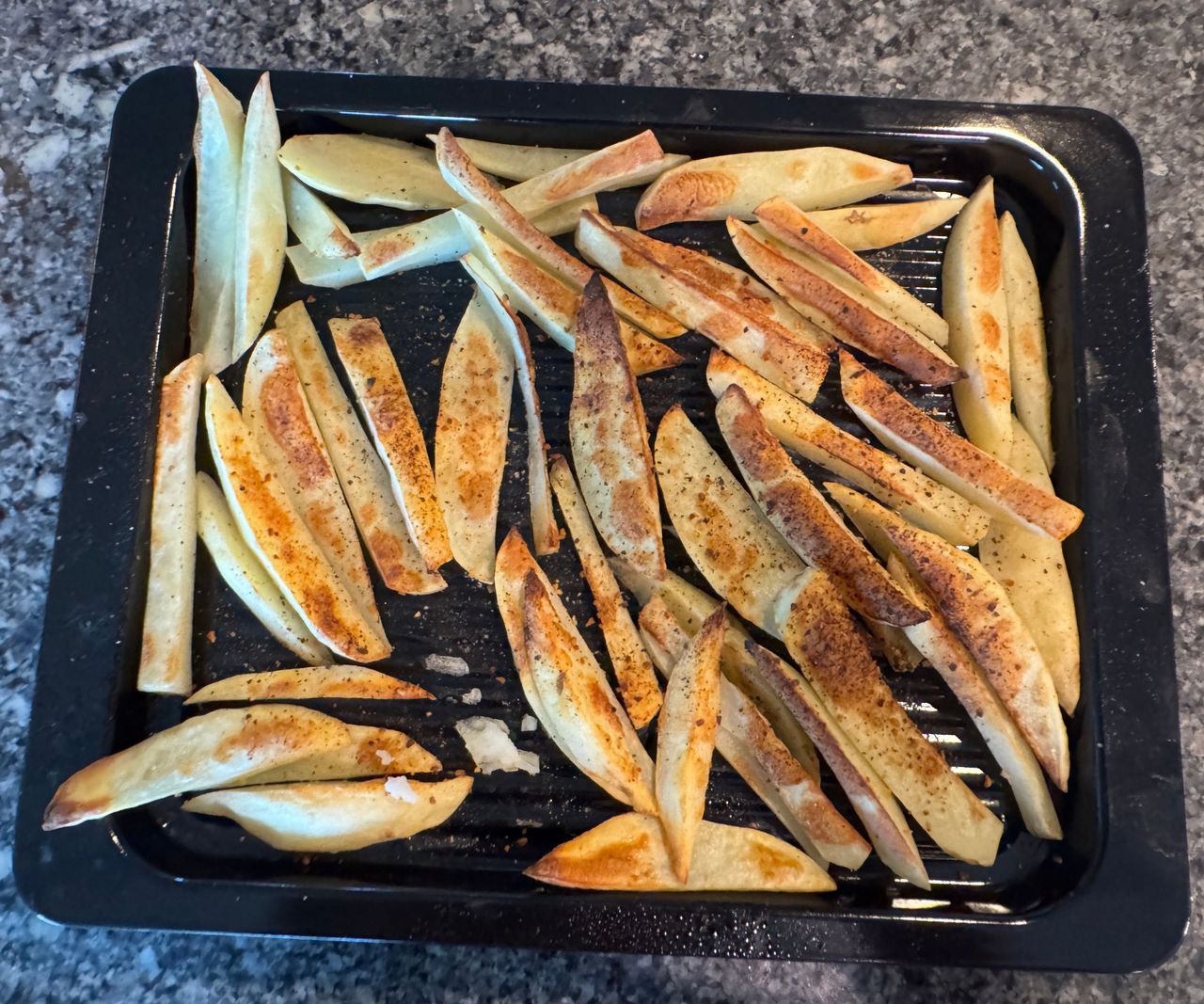 A black baking tray of lightly fried potato fry slices