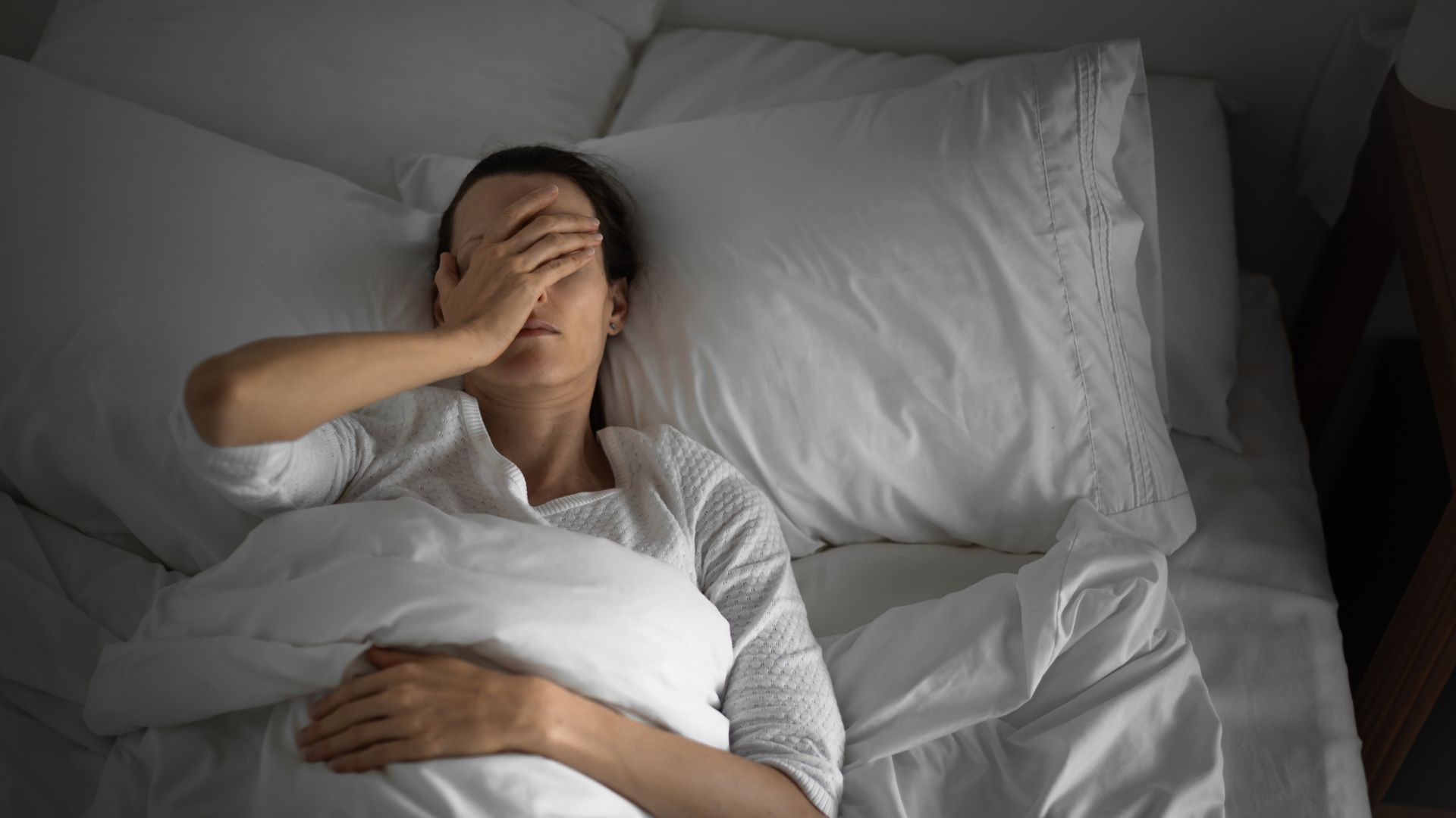 A woman lies on her back in a bed that has white bedding, covering her face with one hand.