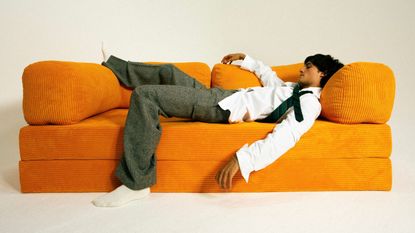 orange OMHU TEDDY sofa with man wearing white shirt, black tie, and gray trousers lying on it