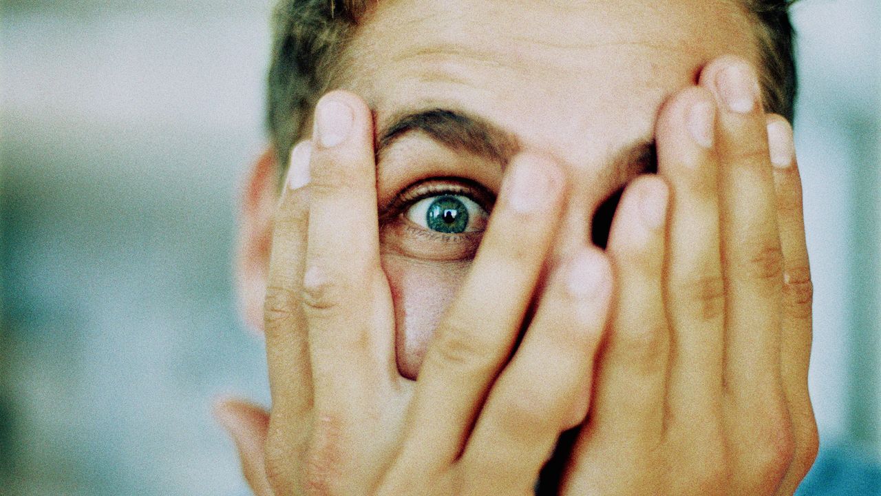 A man has his hands over his face and looks scared as he peeks through his fingers.