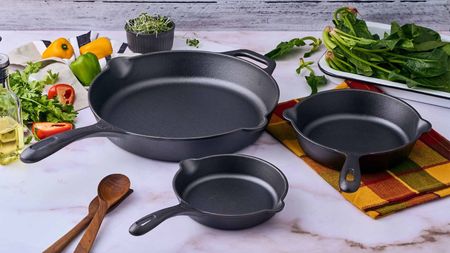 Three black cast iron skillets on a marble worktop surrounded by fresh produce. 