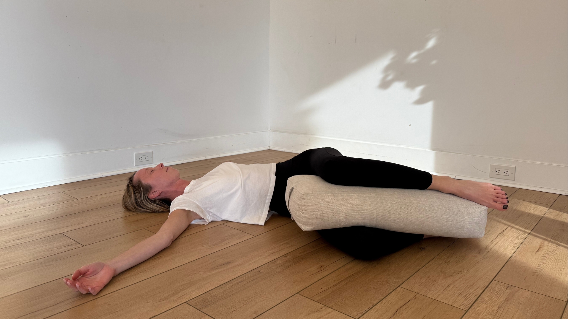 woman lying on her back with knees bent to one side in a spinal twist with a large cushion in between her knees, on a wooden floor with white walls behind her