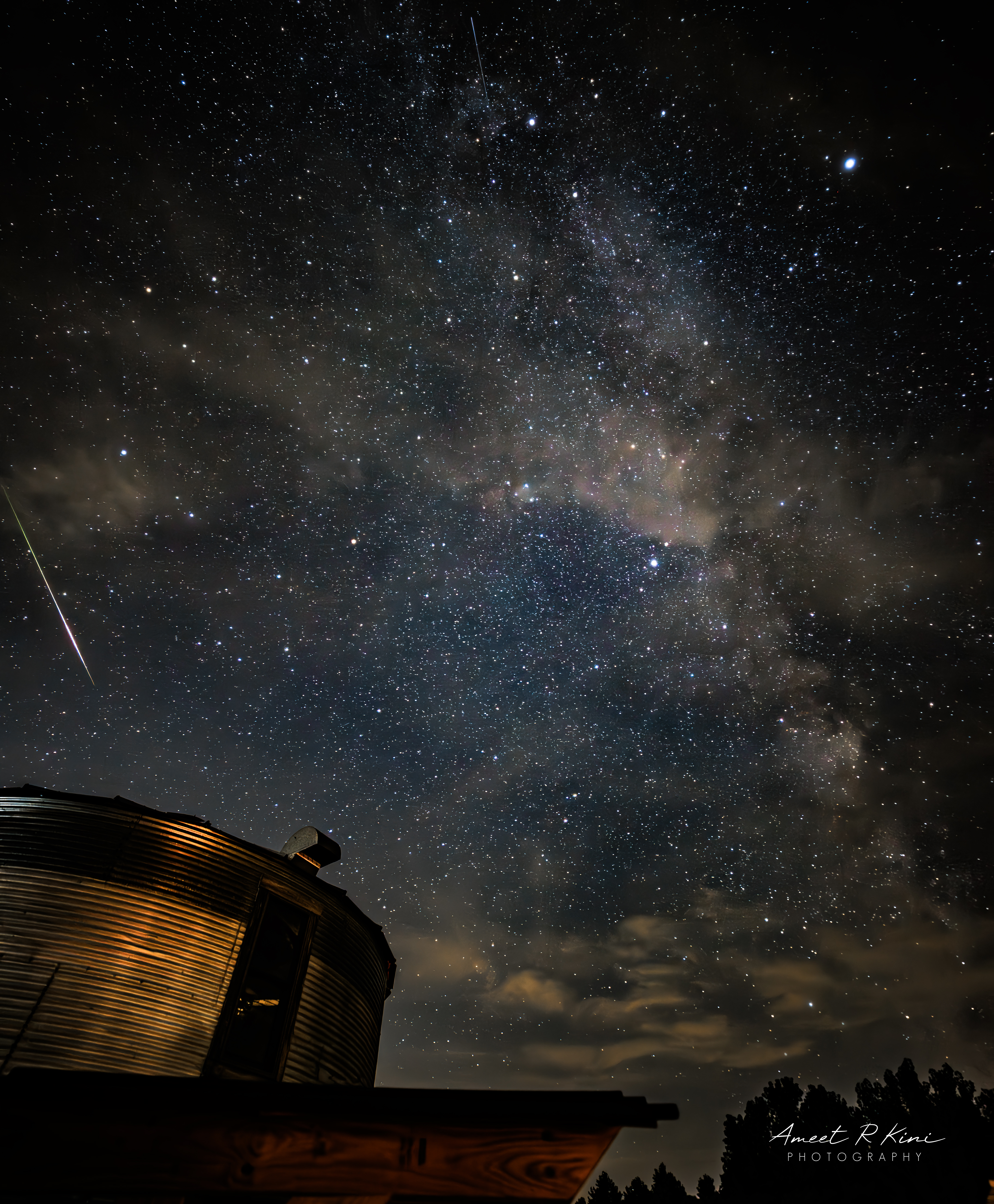 A shooting star is pictured streaking through the night sky alongside the glowing band of the Milky Way above a metal grain silo.