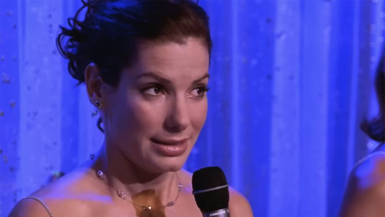 Gracie Hart, AKA Sandra Bullock, speaking onstage in the middle of Miss Congeniality pageant. She is holding a microphone and standing in front of a blue stage backdrop.