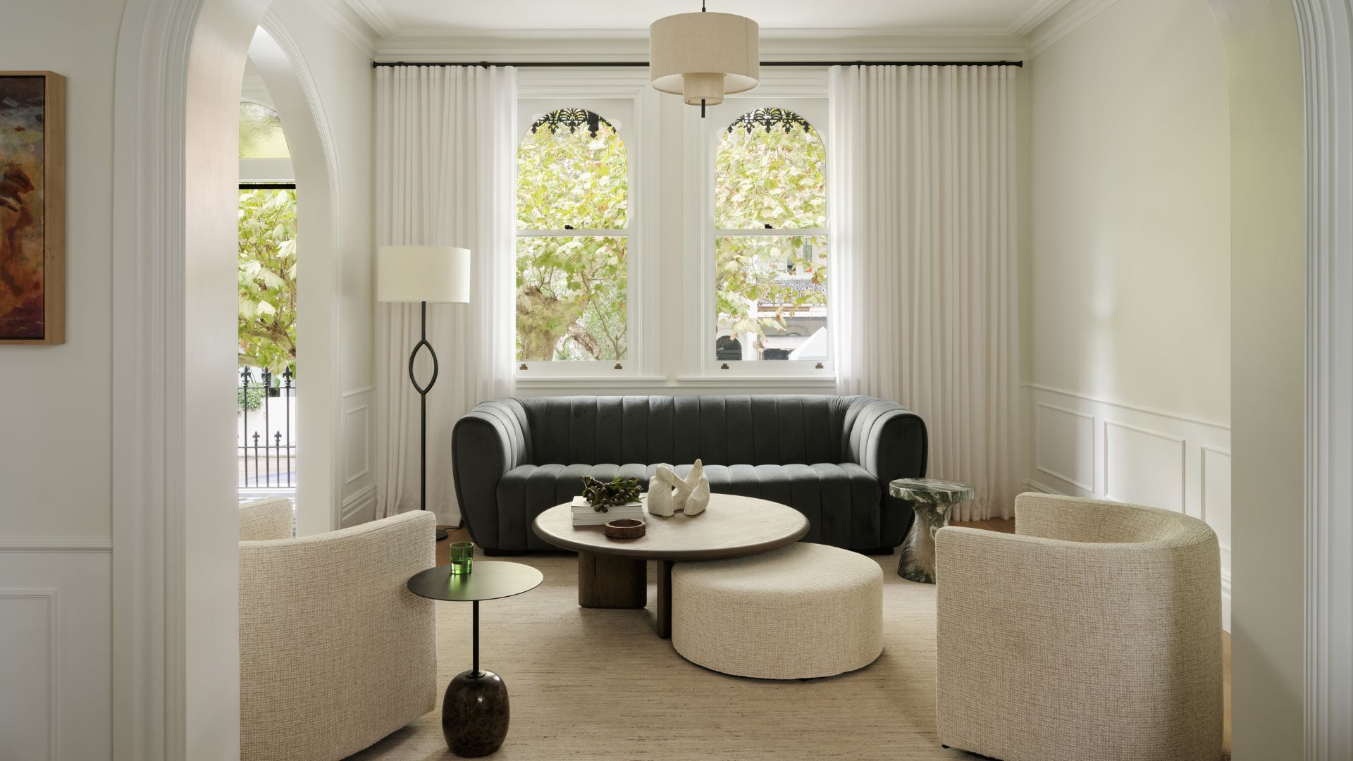 A bright white living room with white curtains, arched windows, a green couch, a pair of white sofa chairs, a coffee table with a white pouf, a standing lamp, and a black side table with a green glass