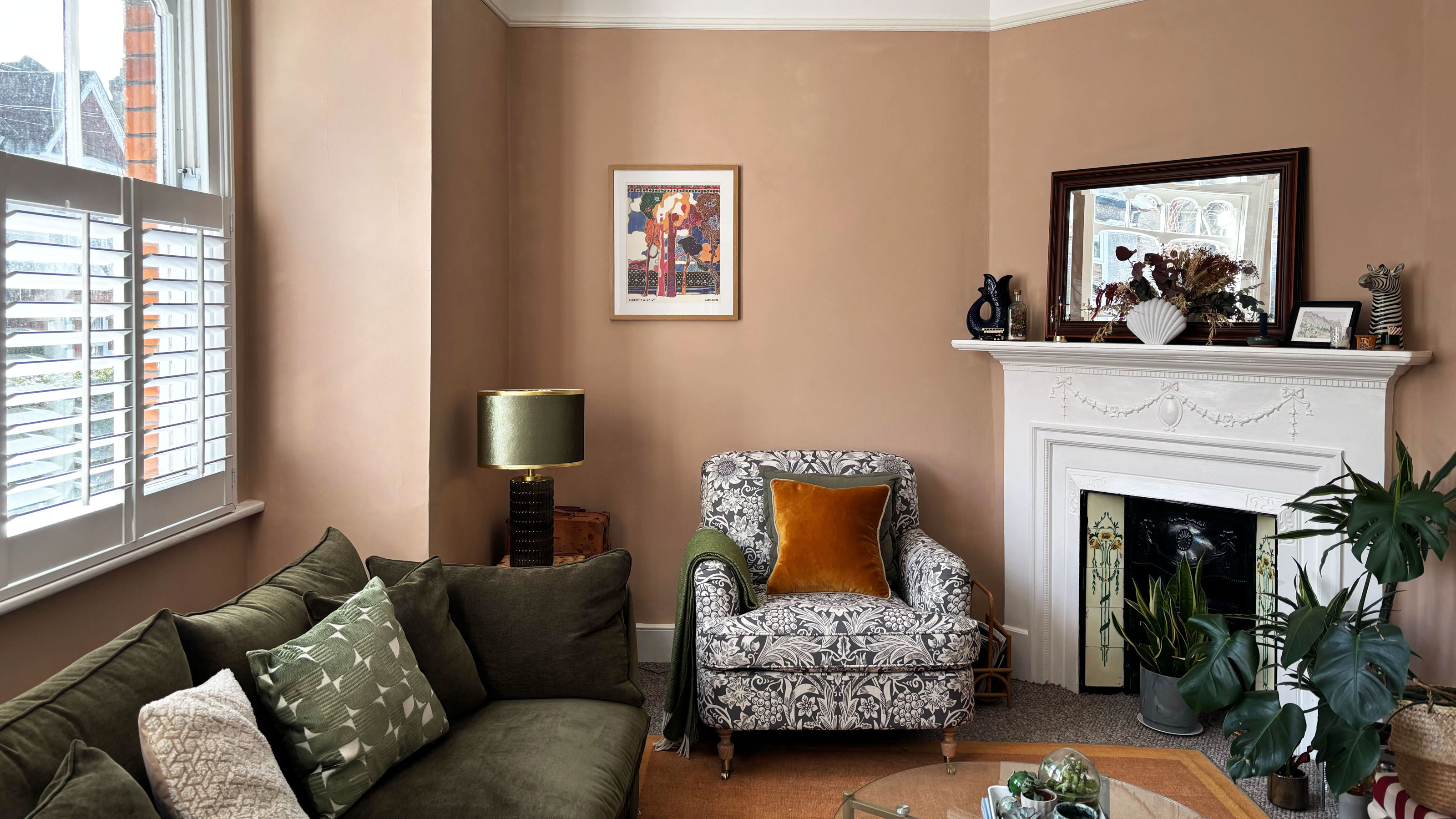 Plaster pink living room with a grey and white patterned armchair, white picture rail and moss green sofa.