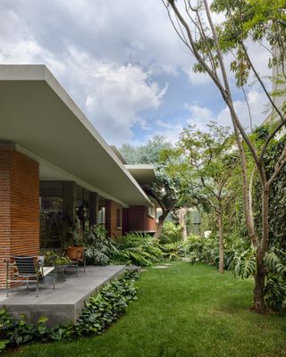 Daytime exterior side image of Ventana House in Mexico among greenery, flat white roof, red brick supporting pillar, green lawn, shrubs, plants and trees, windows, stone patio area with irregular shaped stone steps, tables and chairs, blue sky with light white clouds