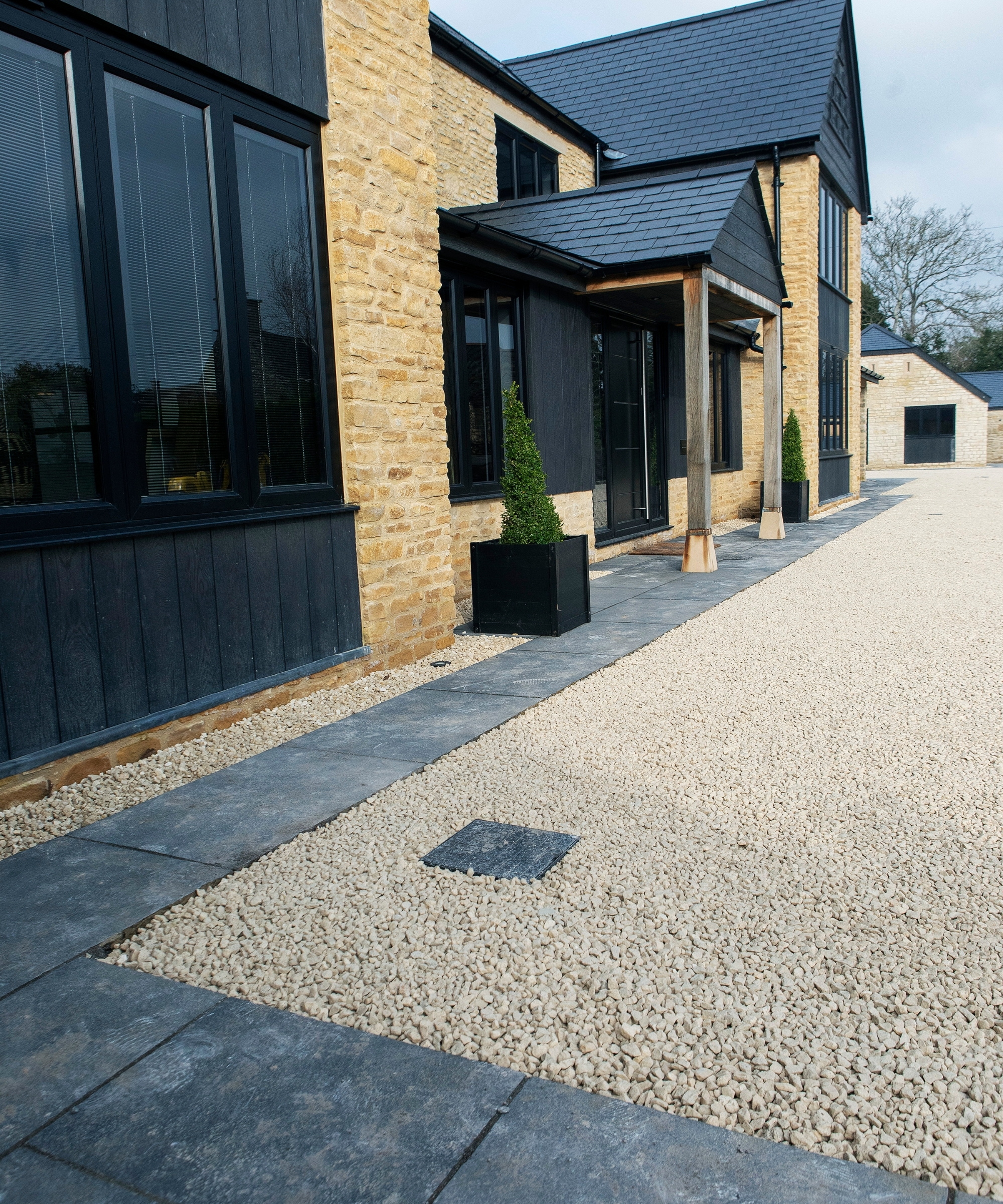 Stone and black-clad house with a slate patio path bordering a pale gravel driveway and topiary planters.
