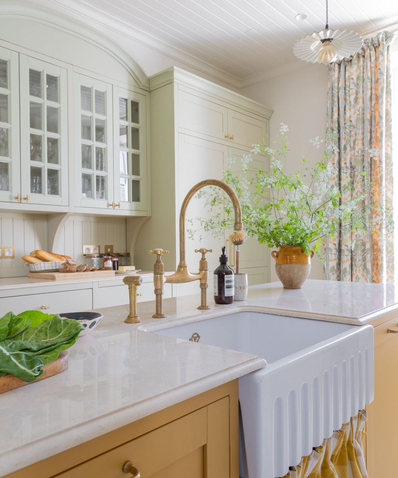 A close-up of a white fluted farmhouse sink with a vintage-style brass bridge faucet on top of a mustard yellow island. Light green glass-front cabinets and a tall floral-patterned curtain are visible in the background