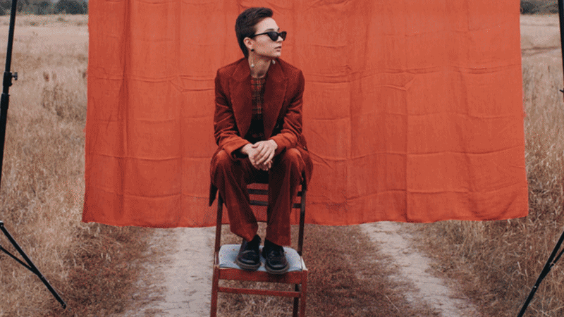 GIF of a person in brown clothing in front of an orange background being transformed into blue clothing and background via Adobe's AI Assistant