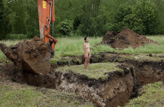 A naked woman stands in the middle of a green field on an island of dirt as a scraper digs a hole all around her.