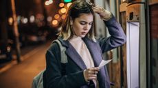 Worried young woman standing in front of an ATM machine and looking at her balance statement