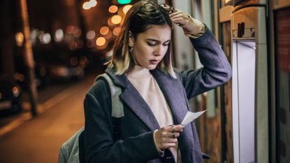 Worried young woman standing in front of an ATM machine and looking at her balance statement