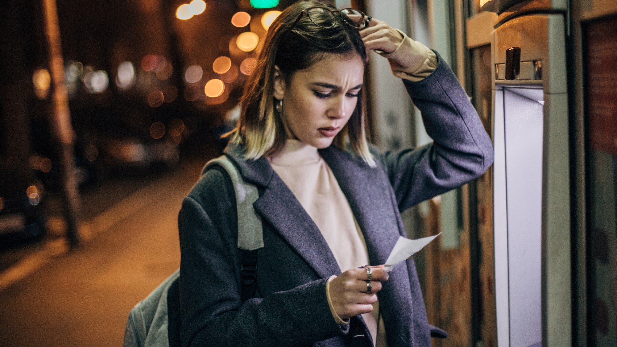 Worried young woman standing in front of an ATM machine and looking at her balance statement