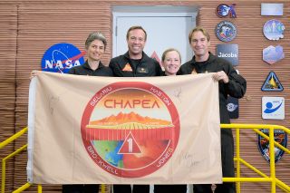 four smiling people holding up a tan banner with the logo of the chapea mock mars mission on it