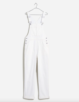 a pair of white overalls hangs on a hanger in front of a plain backdrop