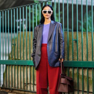 Pornwika Spiecker wears gold earrings, white sunglasses, a purple ribbed t-shirt, a black shiny leather long blazer jacket, high waist red large pants, a burgundy shiny leather large handbag, black and white leather pointed pumps heels shoes, outside the Stella McCartney show, during Paris Fashion Week - Womenswear Spring Summer 2022, on October 04, 2021 in Paris, France.