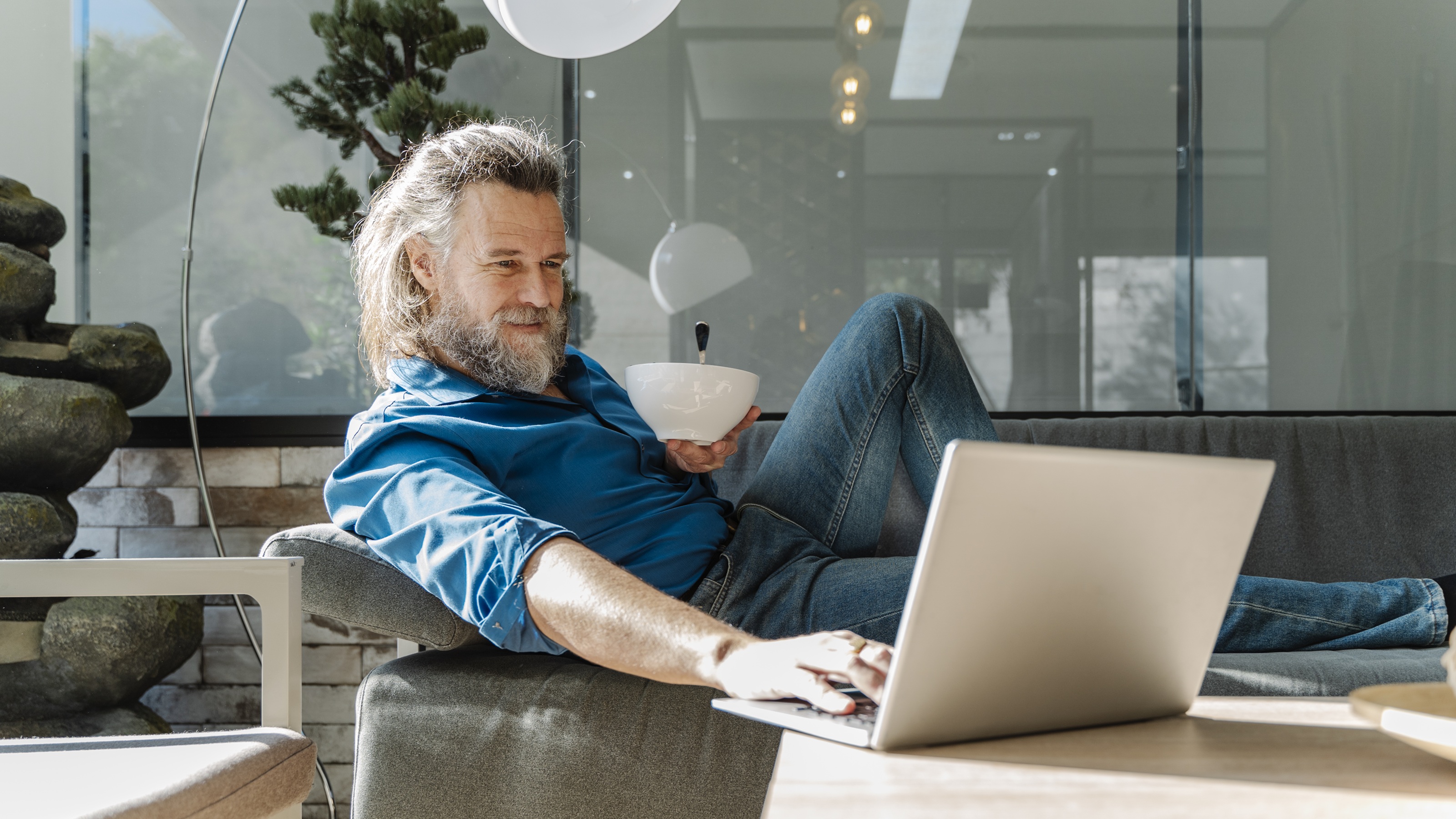 A smiling older man relaxes on the sofa and looks at his laptop.