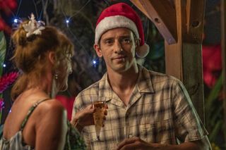 A still from the Death In Paradise Christmas Special showing Neville (Ralf Little) standing at a Christmas party wearing a Santa hat and a casual shirt, holding a glass of bubbly. Melanie (Doon Mackichan) is talking to him, standing with her back to the camera.