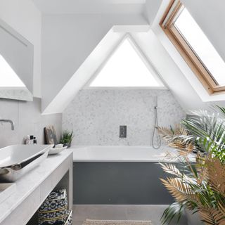 A loft bathroom, sloping eaves and skylight, a bath with grey tiles and silver coloured basin and mat
