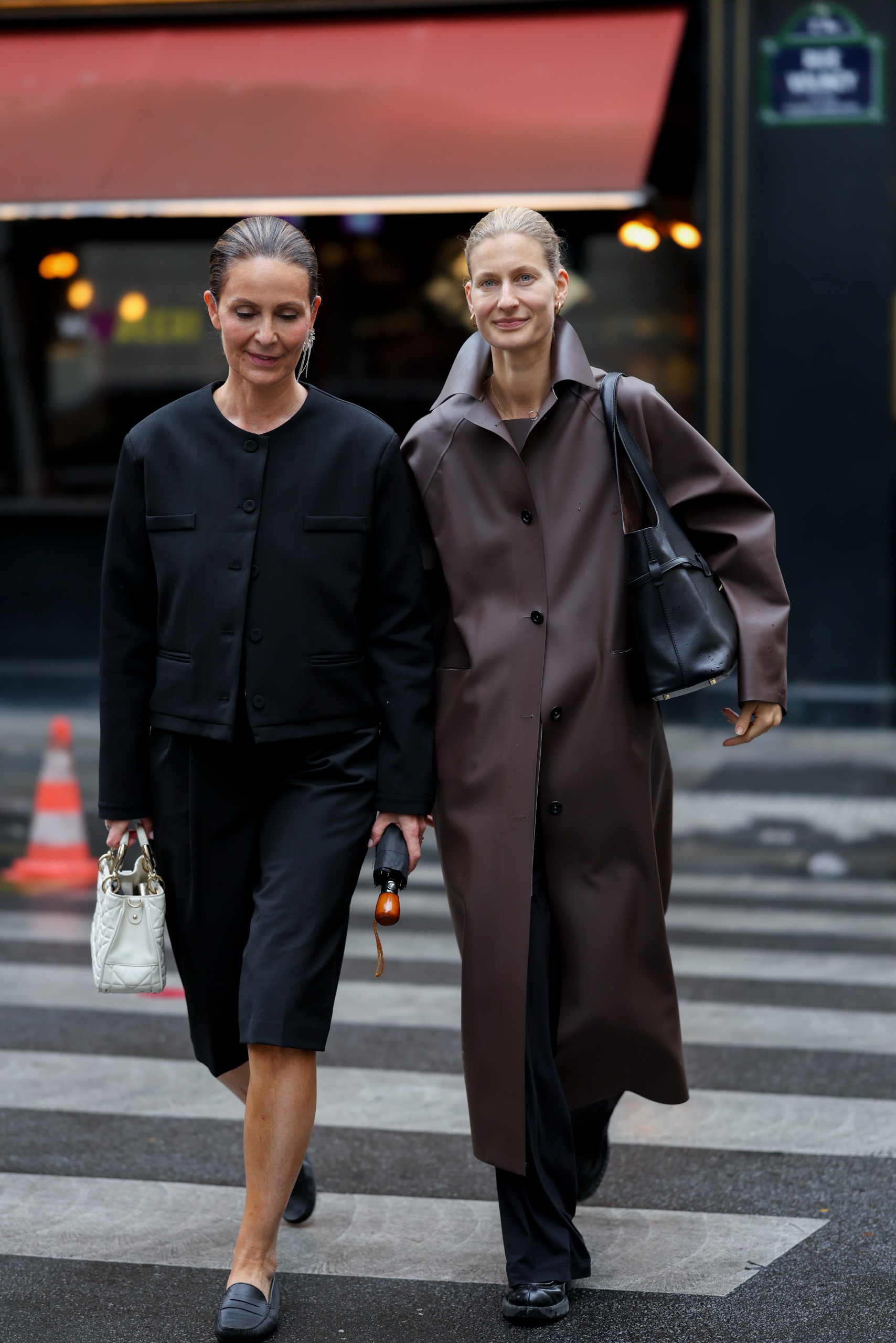 Miluska Van't Lamnwearing a black collarless jacket; black tailored shorts; leather loafers; a white Dior bag; Simone Sniekers is wearing a brown long coat; black wide leg trousers; chunky loafers
