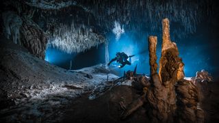 A diver explores an illuminated underwater cave, surrounded by stalagmites and stalactites with blue lighting