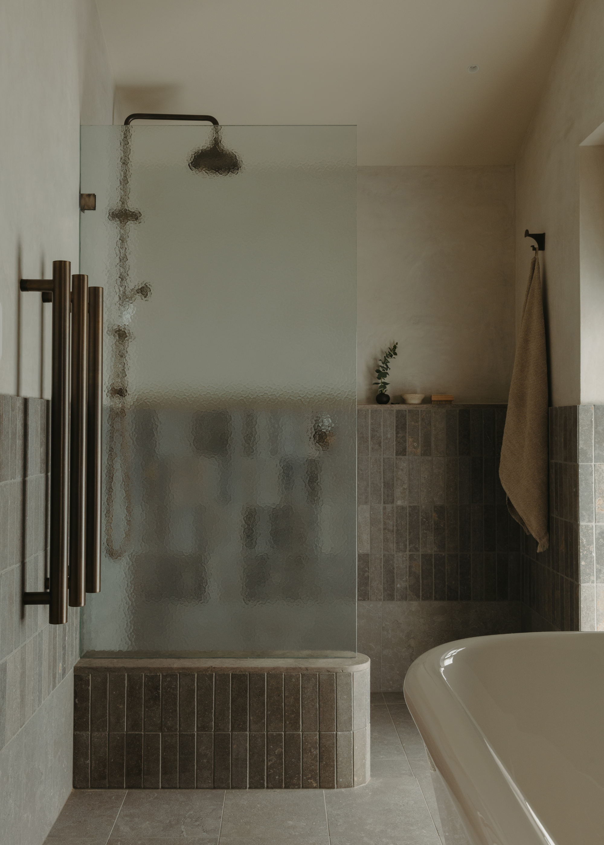 A dim bathroom with a tub, and a shower space with black hardware, frosted glass, and a tiled wall