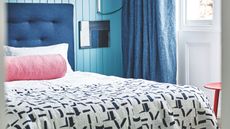 Blue panelled bedroom with blue double bed with white bedding, pink cylindrical cushion, patterned quilt, and large white windows with dark blue curtains beside a red stool.