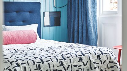 Blue panelled bedroom with blue double bed with white bedding, pink cylindrical cushion, patterned quilt, and large white windows with dark blue curtains beside a red stool.