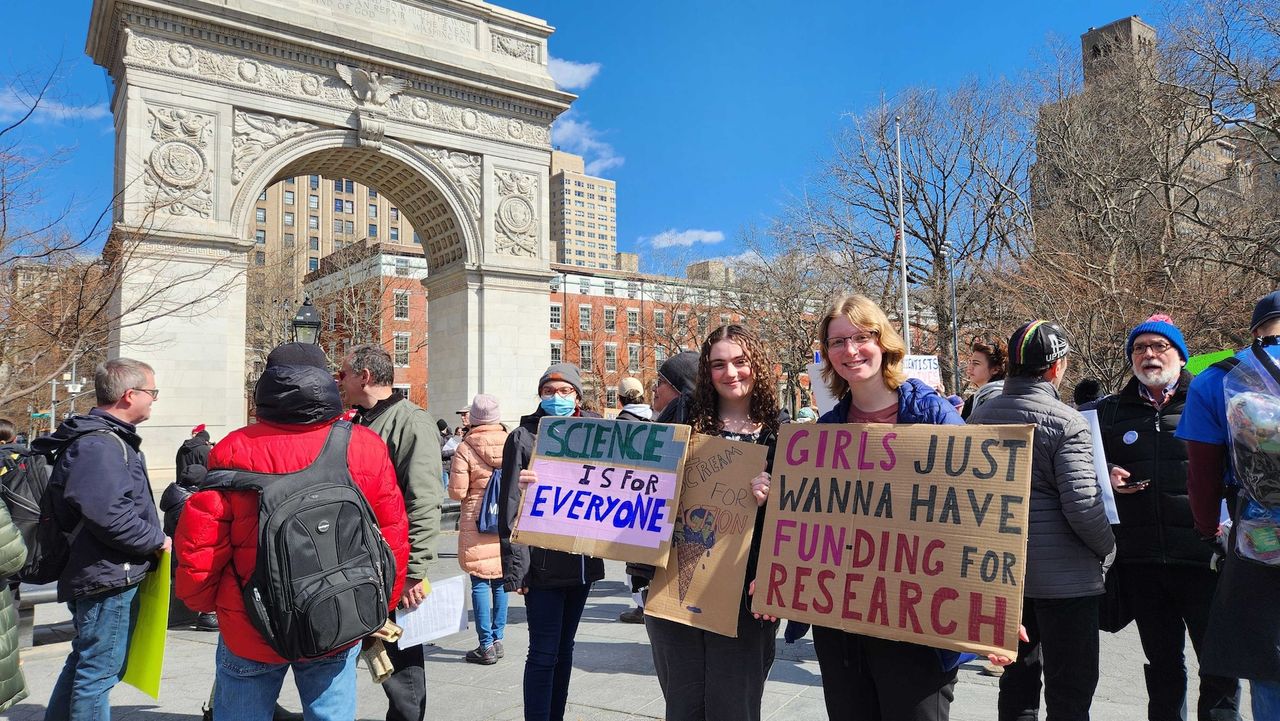 A group of women the Stand Up For Science rally hold protest signs. One says "Science is for everyone" and the other says "Girls just wanna have fun-ding for research".