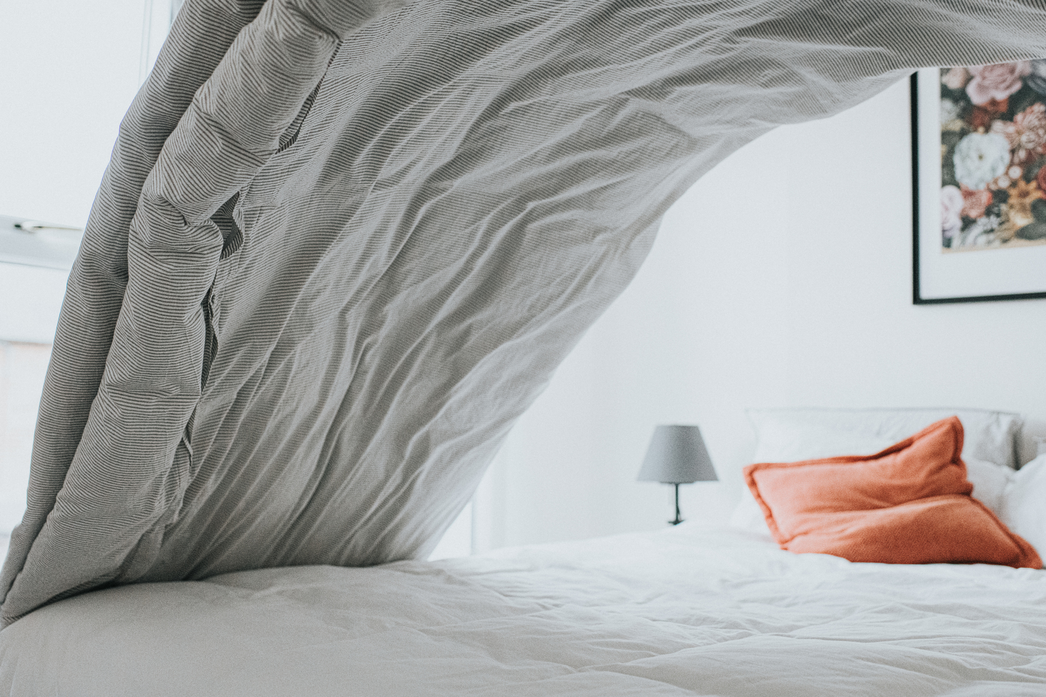 A bedroom with white bed sheets and grey duvet being lifted in air, table lamp with grey lampshade and orange scatter cushion