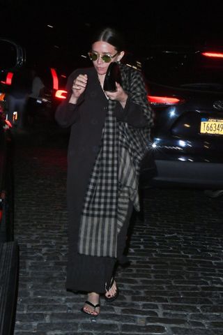 Ashley Olsen wearing flip-flops with a long black coat, sunglasses, a checkered scarf, and a black clutch.