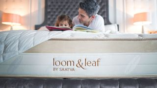 Saatva Loom & Leaf mattress image shows a man and his daughter read a book together while lying on the Loom & Leaf mattress topped with a grey button headboard