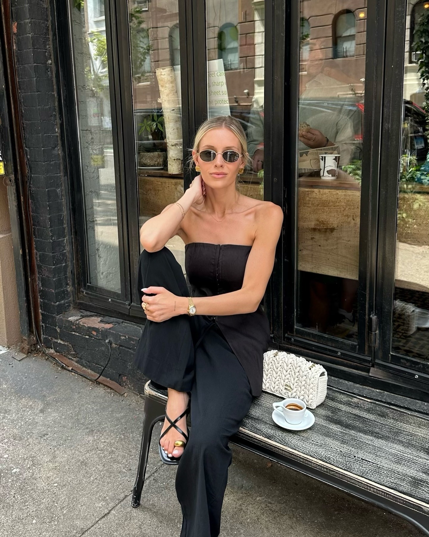 Influencer wears metal detail sandals with black trousers and a black strapless top. She sits on a bench outside of a coffee shop.