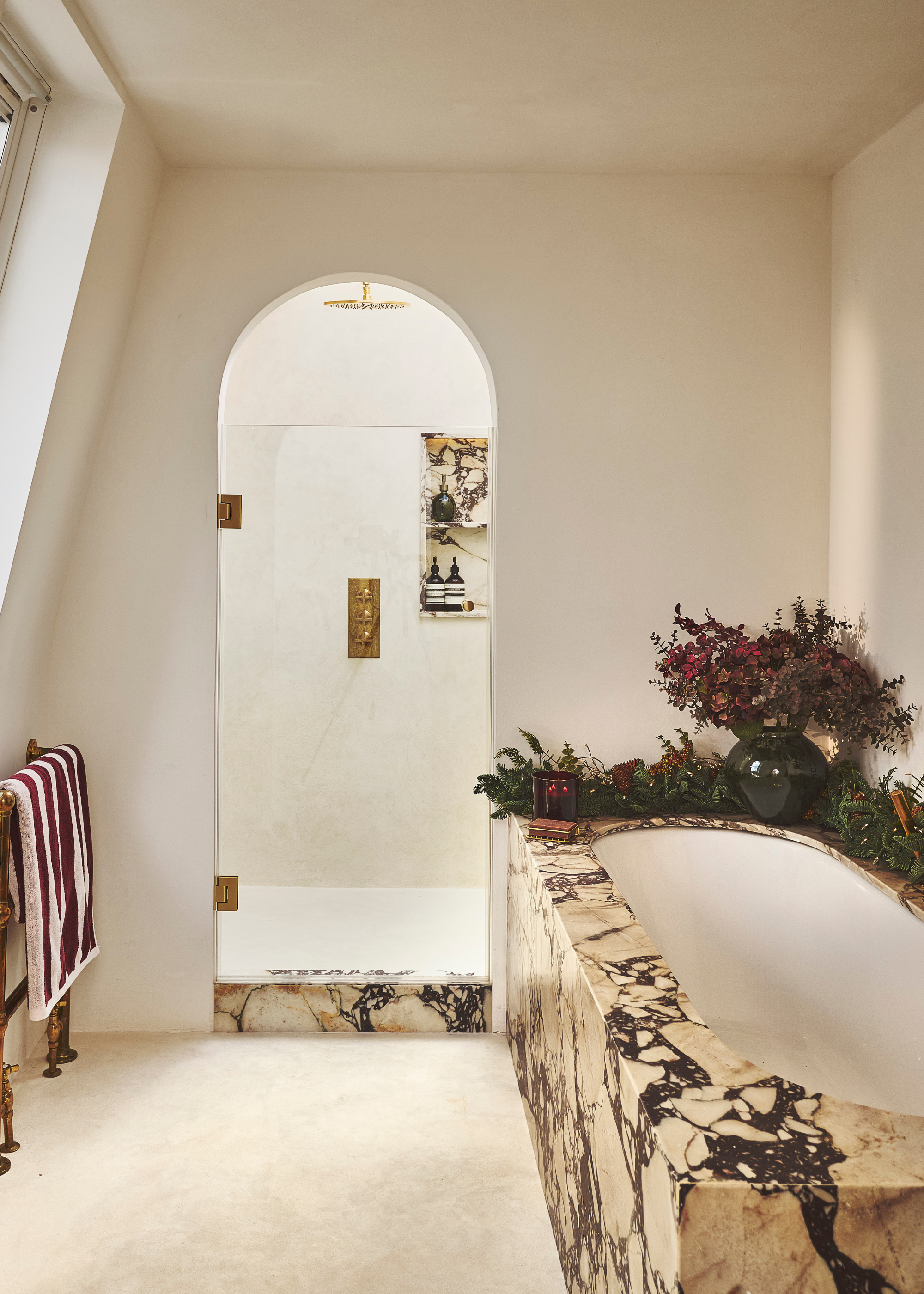 A modern marble bathroom decorated for christmas with a fresh garland around the bathtub and florals in a vase with a glass arched door leading to a shower area in the background