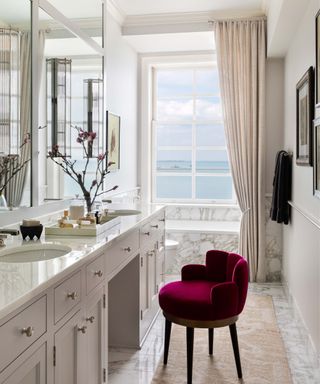 A light-filled bathroom with an outdoor beach view, a marble bathtub, a velvet maroon chair and a vintage runner