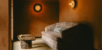 A golden-hued hotel lobby is filled with textural fabrics through a vividly patterned, vintage-looking sofa, styled alongside a wooden coffee table and round, industrial sconces.