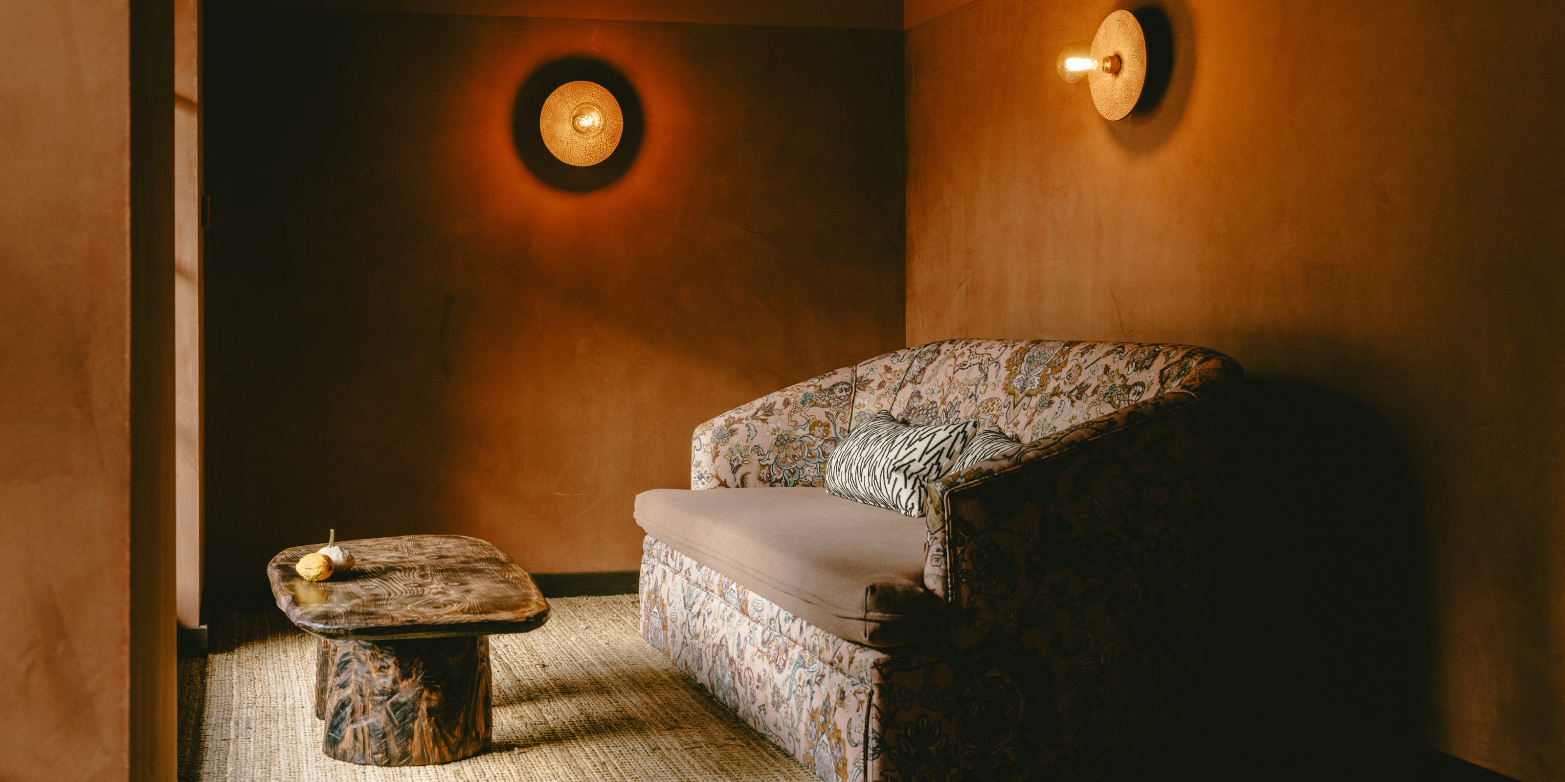 A golden-hued hotel lobby is filled with textural fabrics through a vividly patterned, vintage-looking sofa, styled alongside a wooden coffee table and round, industrial sconces.