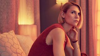 A woman sits on top of a bed with her hand held underneath her jaw